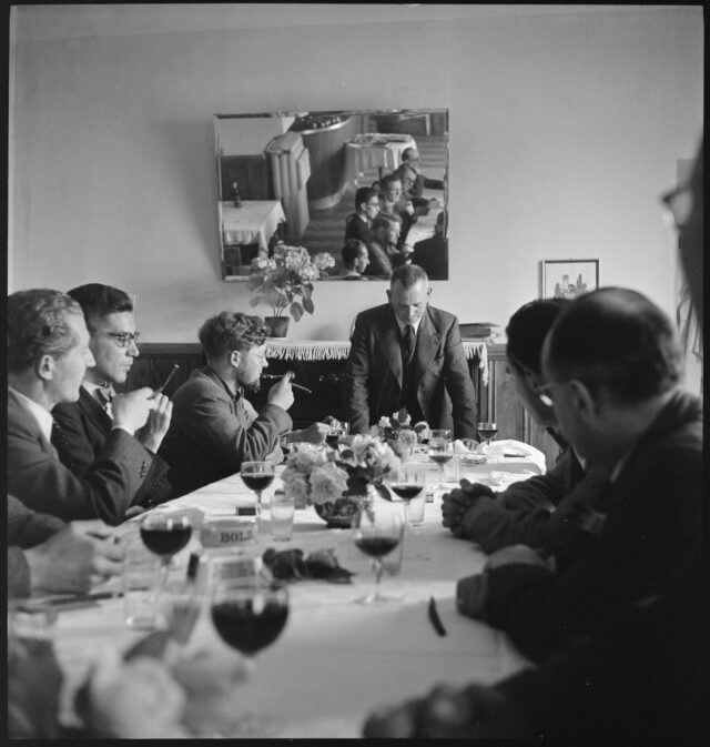 R740: Männer sitzen, teils rauchend, um einen dekorierten Tisch