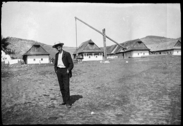 Janko Cadra debout devant trois maisons et un puit [?] dans la campagne slovaque