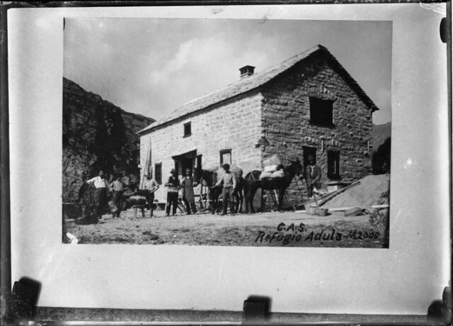 Fotografia di una fotografia (DON2896): gruppo di uomini con cavalli davanti alla capanna dell'Adula; quarto da sinistra: il guardiano Innocente Bassi