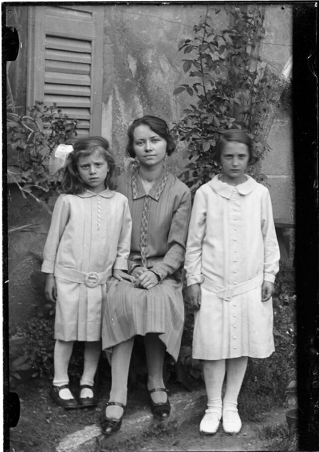 Irene Belgeri seduta tra due bambine vestite di bianco davanti a una casa