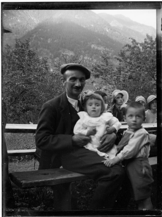 Mario Bozzini con due figli seduti a un tavolo all'aperto; sullo sfondo un gruppo di giovani donne