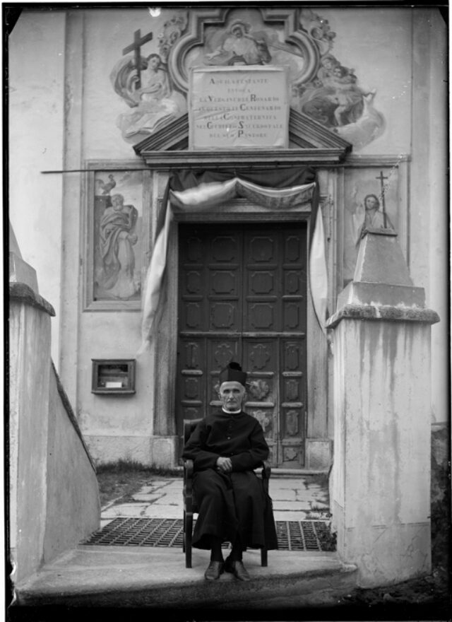 Giacomo Valchera, sacerdote, seduto davanti alla chiesa parrocchiale di Aquila