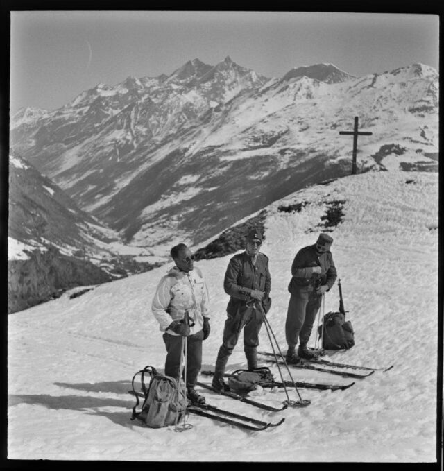 [Drei Skifahrer nach Aufstieg auf Berg vor Bergpanorama]
