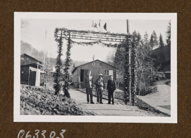 Drei Soldaten unter bekränztem Eingangstor (Beschriftung: III Bat, RKP32 (?)), im Hintergrund Gebäude