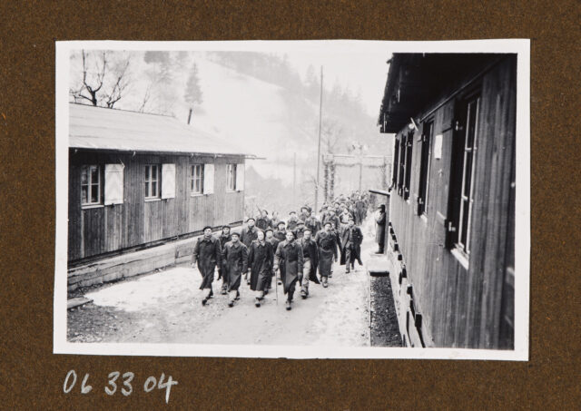 Soldaten (Internierte?) in Mänteln zwischen zwei Gebäuden