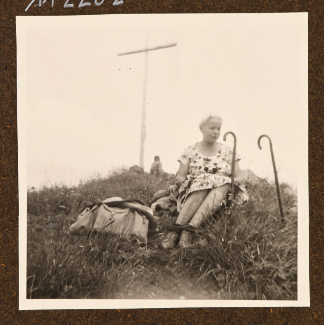 Bettina Zweifel vor Gipfelkreuz mit zwei Spazierstöcken im Gras sitzend