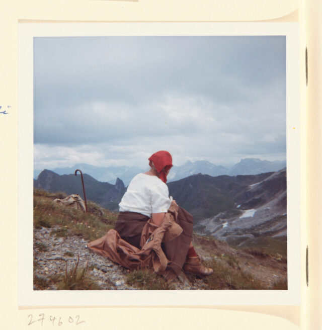 Bettina Zweifel mit rotem Kopftuch (Rückenansicht) sitzend vor Bergpanorama