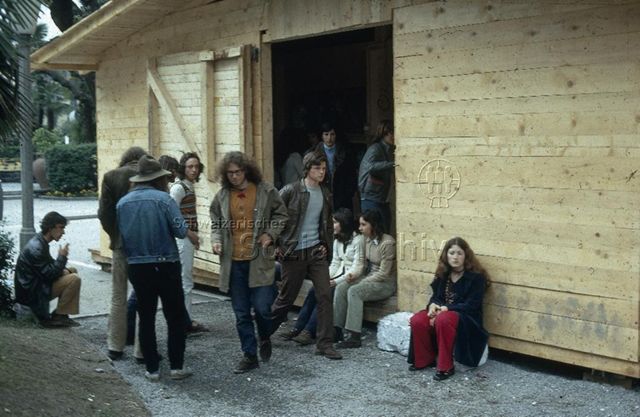 "Freizeitanlagen, Gemeinschaftszentren: Cantiere della Gioventù, Tessin" - Jugendliche sitzen und stehen vor dem Eingang eines Holzhauses; um 1975