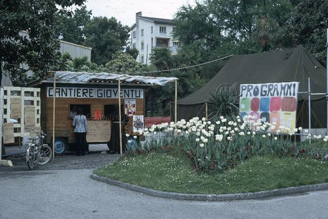 "Freizeitanlagen, Gemeinschaftszentren: Cantiere della Gioventù, Tessin" - Ein hölzerner Verkaufswagen und ein Zelt aus Blache, daran ist ein Transparent befestigt, auf dem farbenfroh das Programm beschrieben ist; um 1975