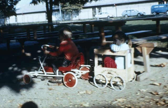 "Freizeitanlagen, Gemeinschaftszentren: FZA Bachwiesen, Zürich" - Zwei Kinder am Spielen, Junge auf Gefährt zieht Mädchen in selbstgebautem Anhänger; um 1965
