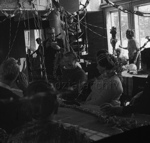 "Freizeitanlagen, Gemeinschaftszentren: Altersnachmittag in der FZA Bachwiesen, Zürich" - Ältere Menschen sitzen um Tische mit Papierschlangen im Haar und auf den Tischen, ein Mann liest aus einem Buch vor; um 1970
