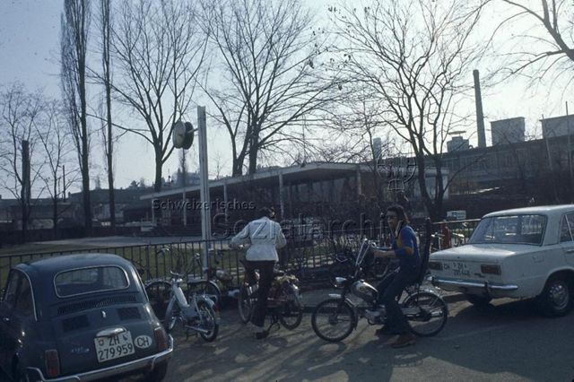 "Freizeitanlagen, Gemeinschaftszentren: FZA Wollishofen, Zürich" - Zwei Jugendliche auf ihren Mopeds, im Hintergrund das FZA mit grossem Hartplatz und Wiese; um 1970