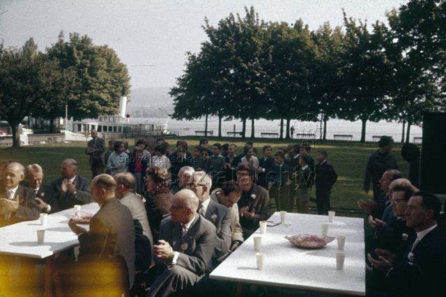 "Freizeitanlagen, Gemeinschaftszentren: Einweihungsfest Wollishofen, Zürich" - Männer und Frauen sitzen applaudierend um Tische im Freien, eine Kindergruppe steht dahinter, im Hintergrund sieht man die Schiffsanlegestelle; 1961