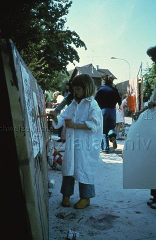 "Freizeitanlagen, Gemeinschaftszentren: Spielaktion Benglen" - Mädchen mit grossem Malhemd zeichnet mit Wasserfarben auf weisses Papier, die Malwand steht im Freien; um 1970