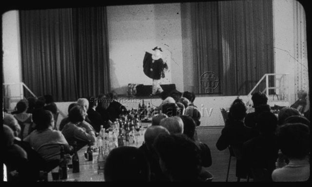 "Kombinierte Gemeindezentren: Schule als offenes Haus, Schulhaus Reute AR" - Theatervorführung, ein Schauspieler steht auf der Bühne mit langen Bühnenvorhängen, an den Tischen sitzen Erwachsene, auf den Tischen stehen Wasser- und Weinflaschen; um 1960