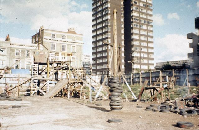 "Europäische Länder: Breakwell Art Center, Milton Keynes GB" - Spielplatz mit aus Holz gezimmerter Burg und alten Autoreifen, das Grundstück ist von einer Holzmauer umgeben, im Hintergrund sieht man Wohnblöcke; um 1960