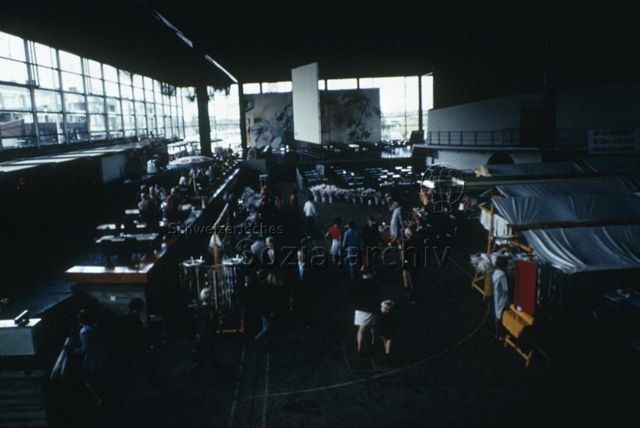 "Europäische Länder: De meer paal, Dronten Holland" - Marktstände in der industriell anmutenden Halle, im Hintergrund steht ein grosses Gemälde, auf einer Galerie stehen Tische; um 1970