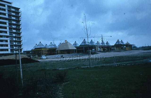 "Europäische Länder: Freizeitzentrum, Eindhoven Holland" - Baustelle von einstöckigen Holzhäusern mit spitzen Dächern auf einer grossen Grünfläche, daneben ein hoher Wohnblock; um 1970