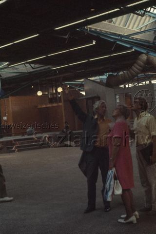 "Europäische Länder: Neusiedlung mit Quartierzentrum, Eindhoven Holland" - Industriell anmutende Halle mit Holzkonstruktionen, ein Mann zeigt Besuchern den Raum; um 1970