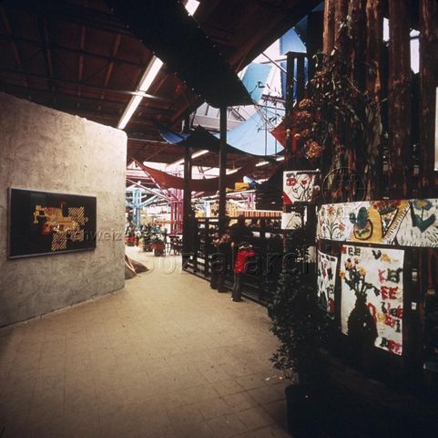 "Europäische Länder: Gemeinschaftszentrum Eindhoven Holland" - Innenbereich, Holzzaun mit Blumen und Zeichnungen verziert, Innenwand mit einem grossformatigen Bild, an der Decke hängen farbige Blachen; um 1970