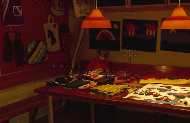 "Europäische Länder: Freizeitzentrum Kopenhagen, Dänemark" - Kleines Mädchen sitzt auf einer Eckbank vor einem Tisch voller unterschiedlicher, farbiger Stoffe, an den Wänden hängen selbstgenähte Gegenstände; um 1970