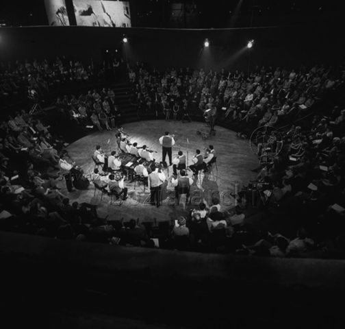 "Europäische Länder: Dronten, Holland" - Eine Musikgruppe spielt in einem Saal, in dem das Publikum kreisförmig und aufsteigend um die Band herum sitzt, das Konzert wird vom Fernsehen gefilmt; um 1970