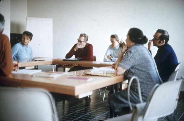 "Freizeitwerkstättenleiter; Tagungen, Kurse: Wochenkurs Herzberg" - Frauen und Männer sitzen an im Quadrat platzierten Tischen mit konzentriertem Gesichtsausdruck, im Hintergrund ein Flipchart; um 1970