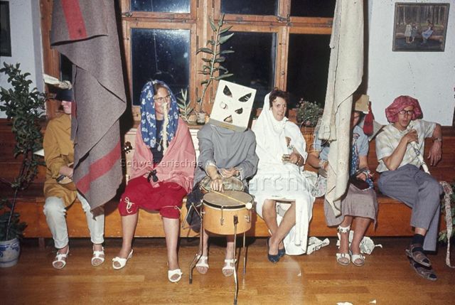 "Spiel- und Sportleiter; Spielwochen und -Kurse: Jeux dramatique, Spielwoche Herzberg" - Verkleidete Erwachsene sitzen auf einer Bank im Innenraum, eine Person hat eine Trommel vor sich und trägt eine Pappschachtel-Maske; um 1970