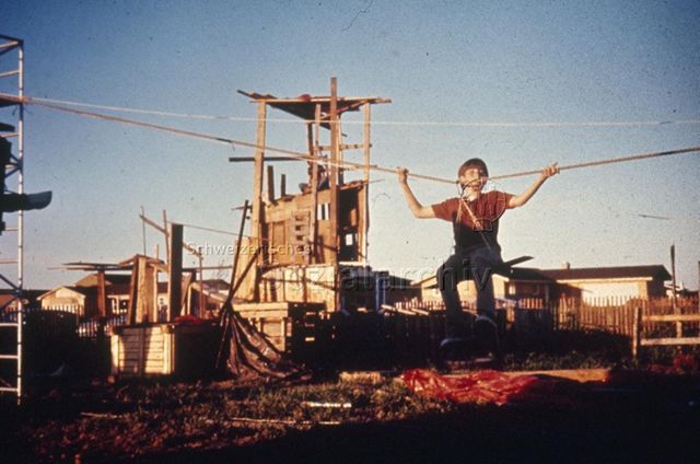 "Ausland; Nordamerika: Champaign Park Distr. Adventure Playgrounds Illinois, USA" - Junge hängt an improvisierter Seilbahn auf Spielplatz mit selbstgezimmertem Holzturm; um 1960