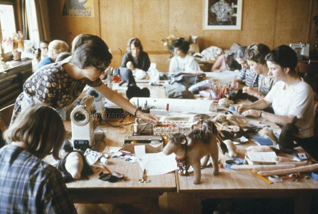 "Freizeitwerkstätten-Leiter: Die Freizeitleiterin Riesbach, Zürich" - Frauen sitzen um einen grossen Tisch mit Nähmaschinen und Nähmaterialien und sind in eigene Näharbeiten vertieft, im Vordergrund steht ein Kamel-Stofftier; um 1960