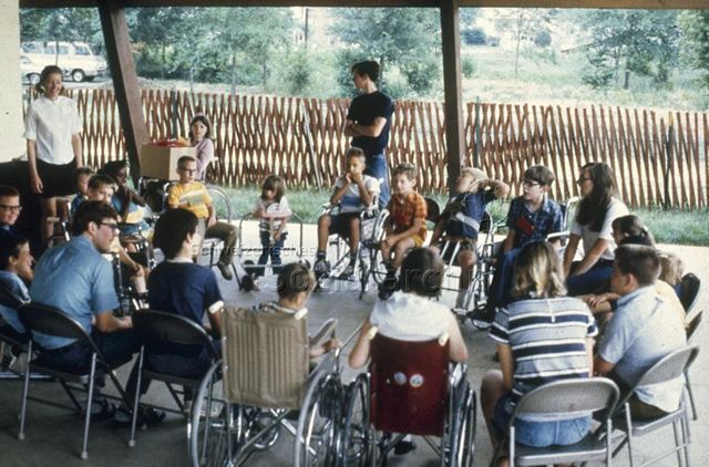 "Ausland; Nordamerika: Handicaped Programm, Champaign Park Distr. Adventure Playgrounds Illinois, USA" - Kinder und Erwachsene sitzen in einem Kreis auf einem Hartplatz im Freien, die Kinder sitzen entweder in einem Rollstuhl oder haben eine Gehhilfe; um 1970