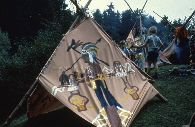 "Spiele im Freien: Denksport, Geschicklichkeit, Benglen ZH, 1976" - Selbstgebaute und -bemalte Tipis am Waldrand, zwei geschminkte Jungen stehen mit Holzstecken neben dem Feuer