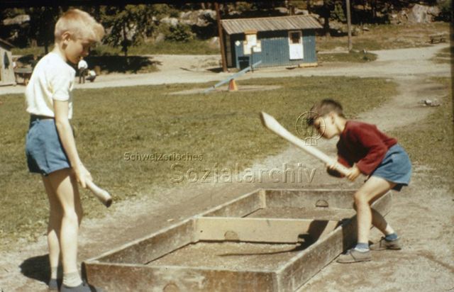 "Spiele im Freien: Ladenhockey" - Zwei Jungen mit Holzknebeln, einer schlägt einen Ball innerhalb eines hölzernen Gestells; um 1960