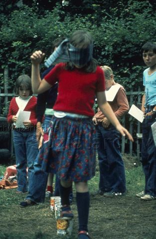 "Spiele im Freien: Robi[nsonspielplatz] in Olten" - Ein Mädchen balanciert mit verbundenen Augen über umgedrehte Konservendosen, andere Kinder stehen wartend in einer Reihe; um 1970