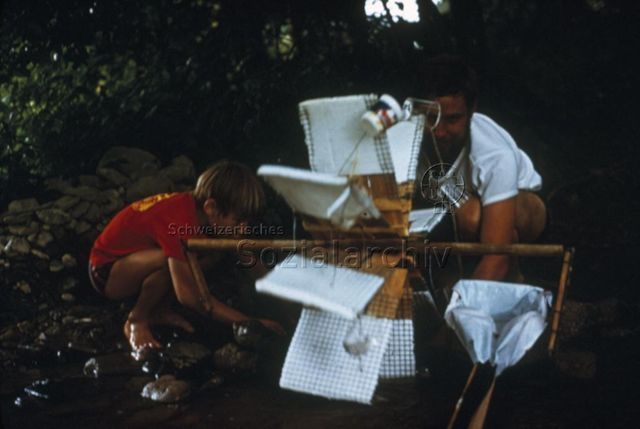 "Spielzeuge aus Holz und Abfällen: Wasserrad" - Ein Junge und ein Mann stehen an einem Bach und bauen ein Wasserrad aus Holz, Draht und Stoff; um 1970