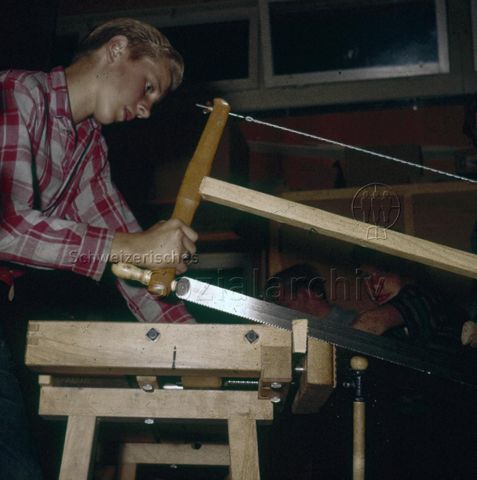 "Schreinern: Mit der grossen Säge" - Junger Mann an Werkbank, ein Stück Holz wegsägend; um 1960