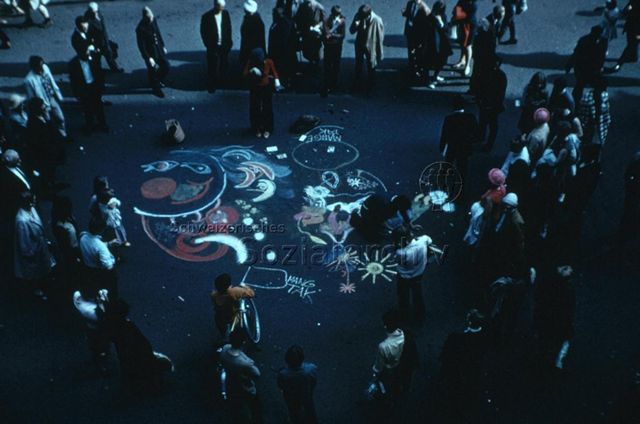 "Zeichnen und Malen: Strassenmalerei" - Eine Frau und ein Mann zeichnen mit farbigen Kreiden auf den Asphalt, zahlreiche Passanten und Passantinnen bilden einen Kreis und betrachten die Arbeit; um 1970
