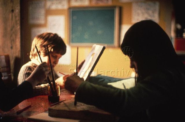 "Zeichnen und Malen: Robi[nsonspielplatz] Allmend, vermutlich Basel" - Kinder sitzen in einem Innenraum am Tisch und malen; um 1970
