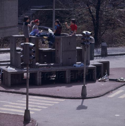 "Verkehrserziehung: Verkehrskindergarten" - Kinder sitzen auf aufeinandergestellten Steinhockern in der Mitte einer Verkehrsinsel mit Strassen und Ampeln in Kindergrösse; um 1970
