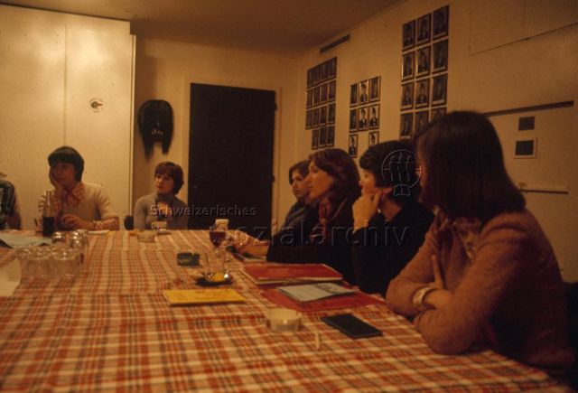 "Elterninitiative: Betreuersitzung Olten" - Frauen sitzen an einem grossen Tisch mit kariertem Tischtuch und hören aufmerksam zu, an den Wänden hängen Fotografien und eine Fasnachts-Maske; um 1970