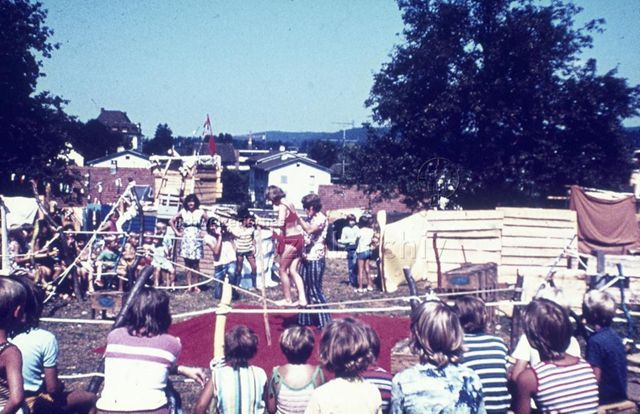 "Kinderzirkus/ Kindertheater: Spielaktion Effretikon ZH" - Ein Mädchen geht über ein an zwei Holzpfählen befestigtes Seil, sie wird dabei gestützt, zahlreiche Kinder schauen , im Kreis rundherumsitzend, zu; um 1970