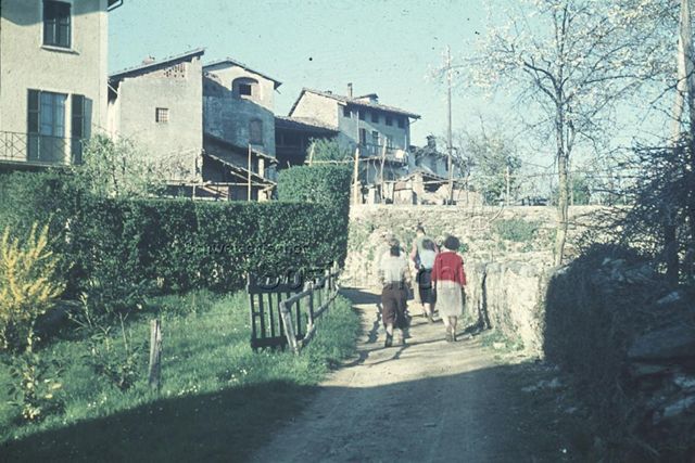 "Wandern: Wanderung im Tessin" - Eine Gruppe Erwachsener geht über eine Naturstrasse in ein Tessiner Dorf hinein; um 1960