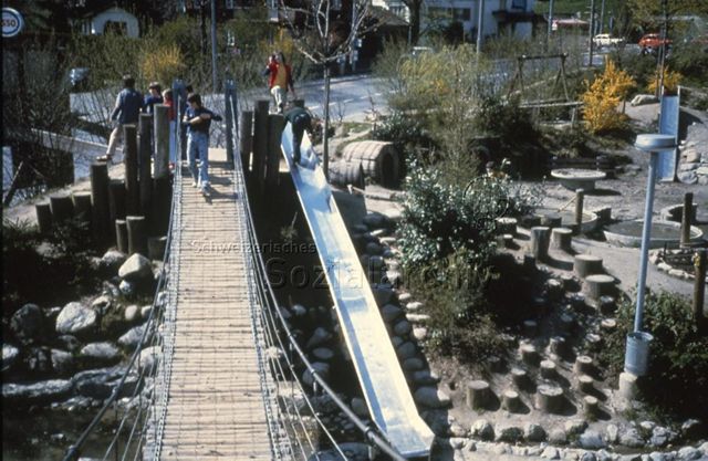 "Siedlungs- und Quartierspielplätze: Spielplatz St. Anton, Luzern" - Spielplatz mit Hängebrücke, langer Rutschbahn, Holzstrünken, Wasserbecken, Kinder am Spielen; um 1970
