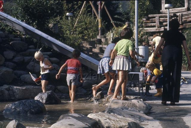 "Siedlungs- und Quartierspielplätze: Spielplatz St. Anton, Luzern, 1975" - Kinder waten barfuss im Wasserbecken neben der Rutschbahn