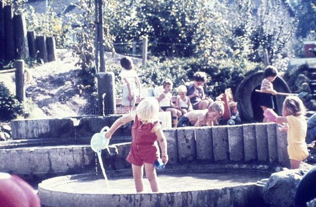 "Siedlungs- und Quartierspielplätze: Spielplatz St. Anton, Luzern" - Ein Kind steht im seichten Wasserbecken und spielt mit einer Giesskanne, im Hintergrund sieht man Kinder im Sandkasten spielen; um 1970