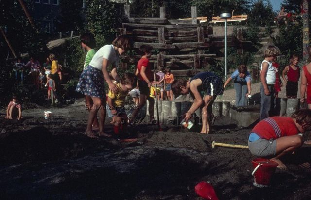 "Siedlungs- und Quartierspielplätze: Spielplatz St. Anton, Luzern" - Kinder spielen mit Wasser im Sandkasten, ein Mädchen hilft einem kleineren Kind; um 1970