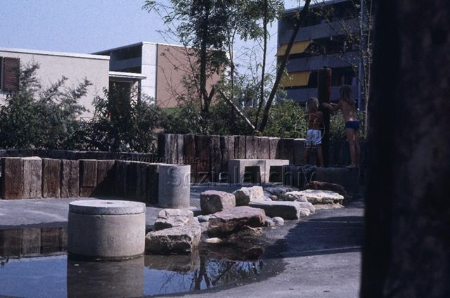 "Siedlungs- und Quartierspielplätze: Augarten Rheinfelden AG" - Zwei Kinder spielen auf einem Spielplatz mit einem kleinen Bach und seichten Becken zwischen Wohngebäuden; um 1970