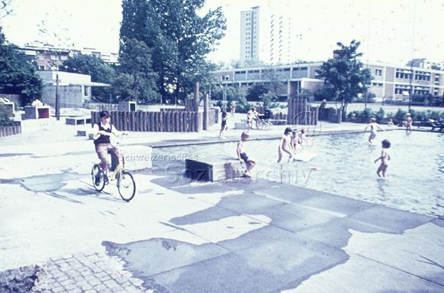 "Siedlungs- und Quartierspielplätze: Siedlungsspielplatz Hirzenbach, Zürich, 1975" - Kinder spielen im einem seichten Bassin, ein Junge fährt auf dem Velo vorbei, im Hintergrund sieht man Wohngebäude