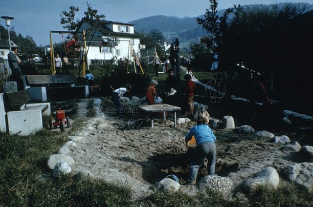 "Siedlungs- und Quartierspielplätze: Siedlungsspielplatz Lichtensteig SG" - Kinder spielen im Sandkasten und auf Schaukeln in einem Park zwischen Wohnhäusern; um 1970