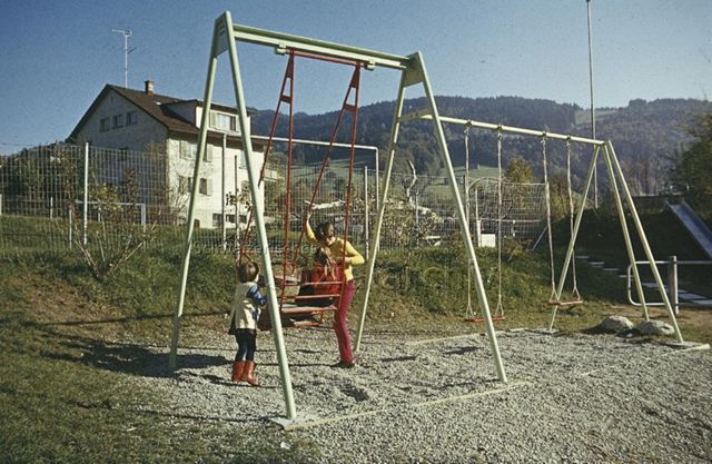 "Siedlungs- und Quartierspielplätze: Kinderspielplatz Lichtensteig SG" - Kinder spielen auf einer Schaukel in einem Park zwischen Wohnhäusern, hinten sieht man einen bewaldeten Hügel; um 1970